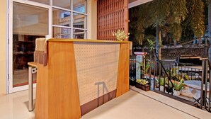 A reception desk with a wooden finish, a glass partition, and a view of a balcony with plants and a car. - OYO 91951 Almira Homestay Syariah (Pekanbaru)