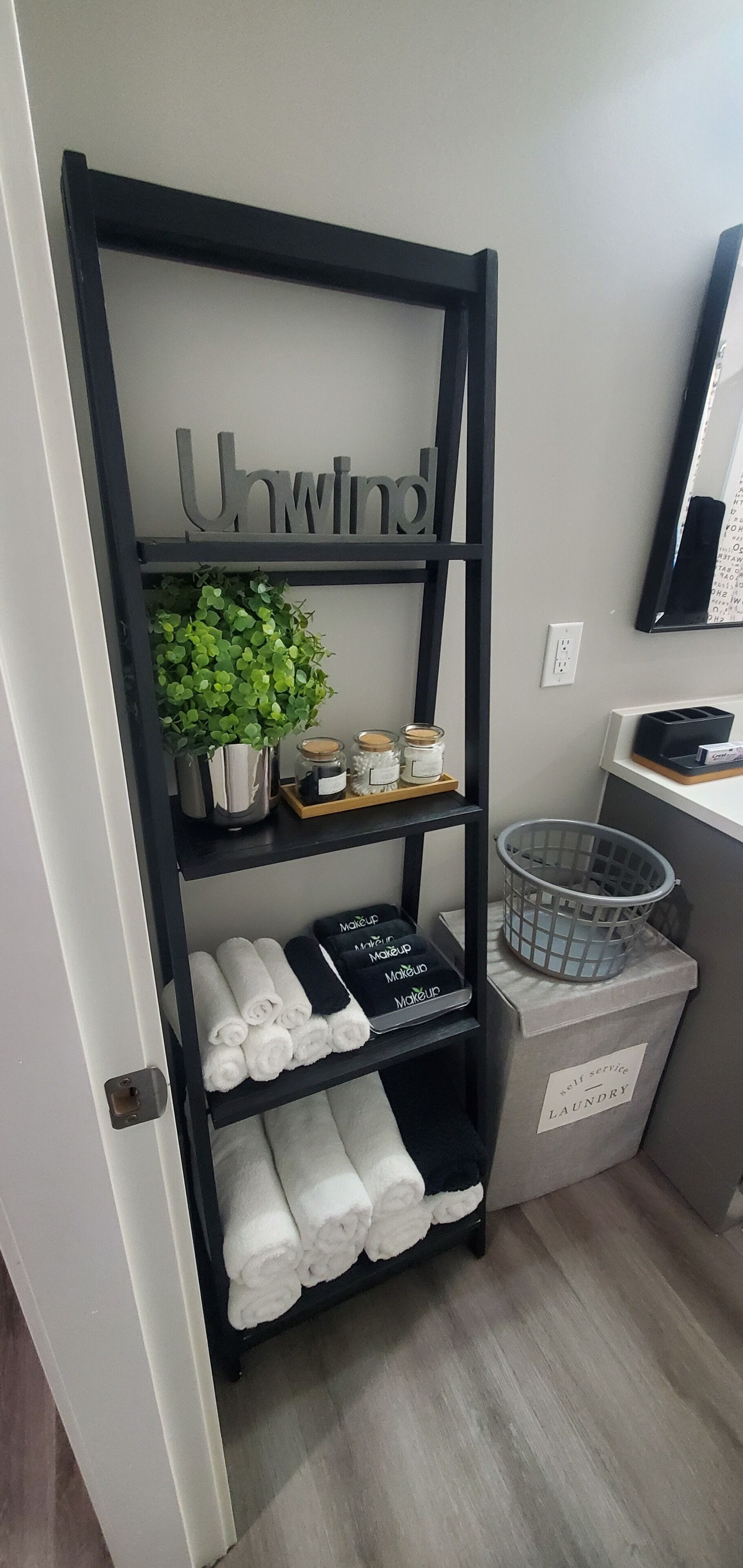 Una estantería de metal negro con un letrero que dice 'Unwind', sosteniendo una planta pequeña, frascos y toallas.