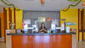 A reception desk with a woman standing behind it, a bulletin board with various notices, and a wall with vertical stripes and a decorative wreath. - Townhouse OAK Hotel Fiducia Pasar Minggu (Jakarta)