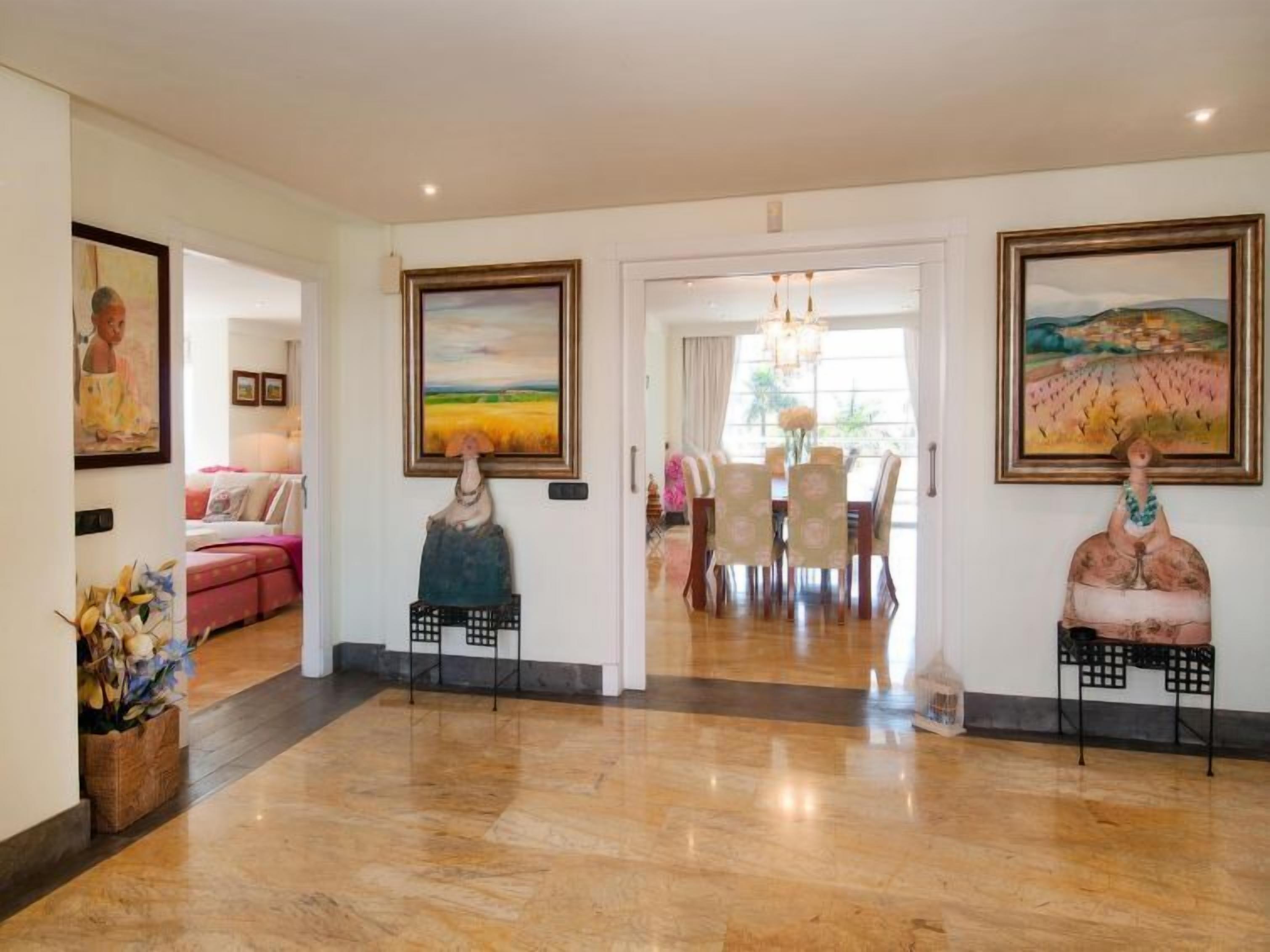 A spacious lobby with a polished wooden floor, a dining area with a chandelier, and two large paintings on the walls.