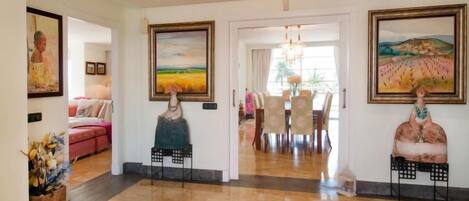 A spacious lobby with a polished wooden floor, a dining area with a chandelier, and two large paintings on the walls.