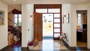 Um corredor amplo com porta de madeira que dá para área externa, escada à esquerda e mesa com cadeira à direita.