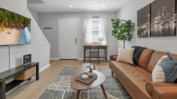 A modern living room with a brown leather sofa, a coffee table, a TV mounted on the wall, and a dining area with a table and chairs. - Urban Townhouse Near Downtown | 2BR + W/D (Columbus)