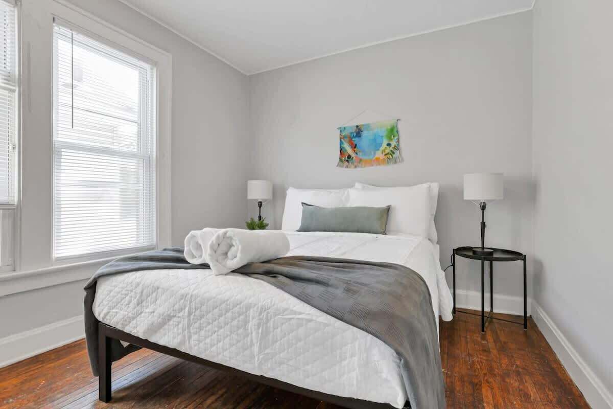 A neatly made bed with a white duvet and gray throw, two bedside lamps, a small plant, and a colorful painting on the wall.