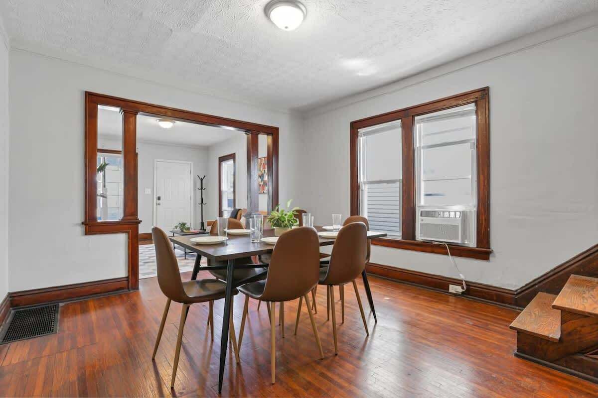 A dining area with a wooden table and four chairs, large windows with wooden frames, and a ceiling light.
