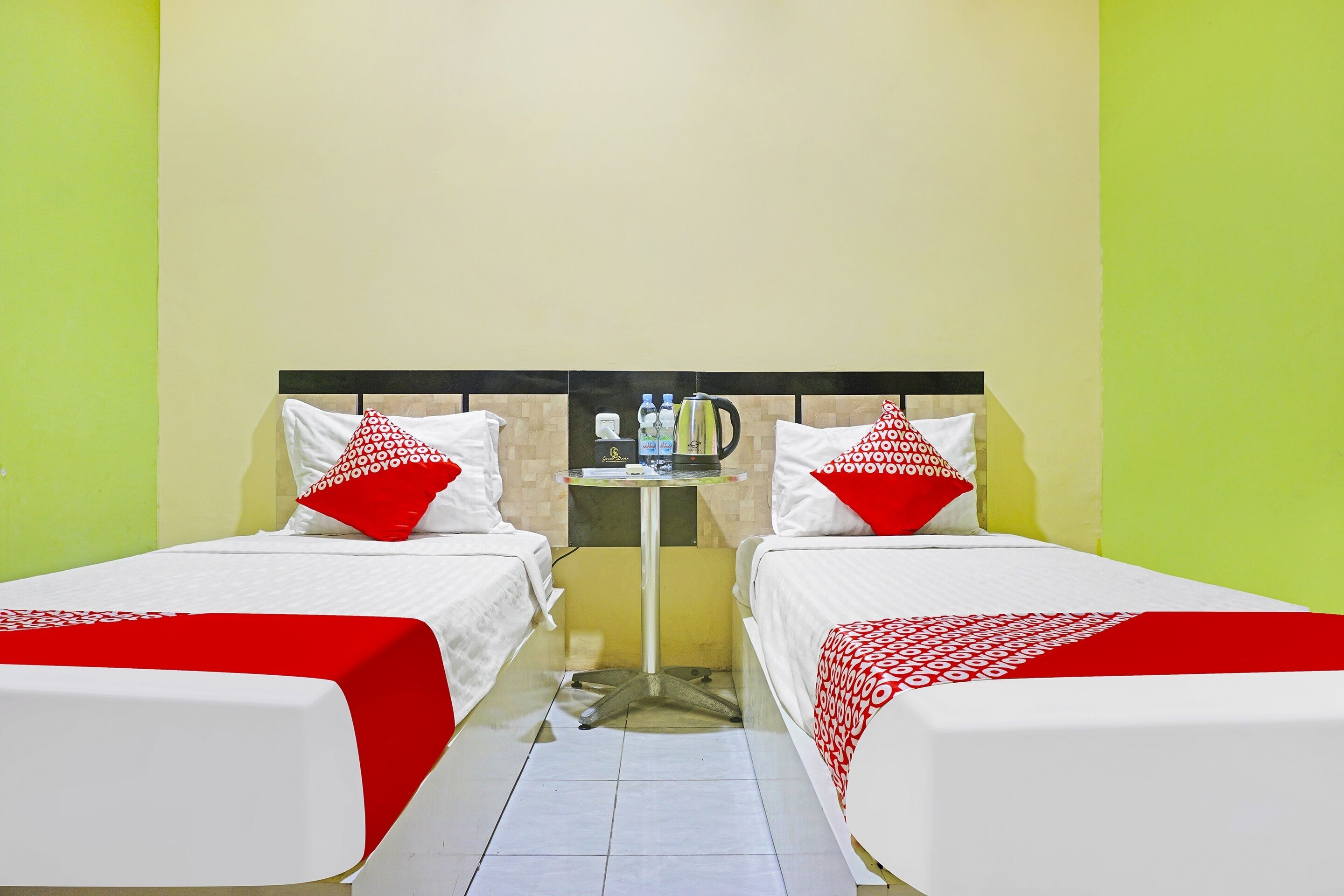 A hotel room with two beds, each with white bedding and red accents, a small table with a kettle and water bottles, and a tiled floor.