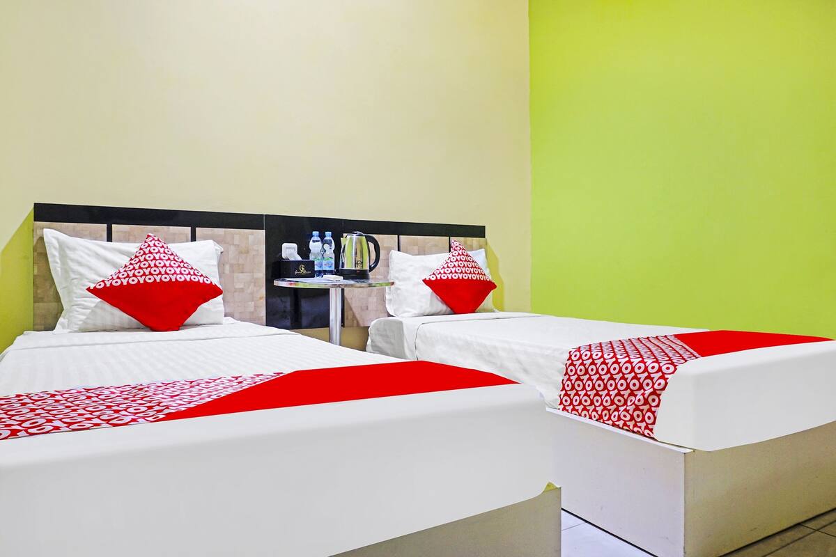 Deux lits simples avec une literie blanche et des touches de rouge, une petite table avec une bouilloire et des bouteilles, et un mur vert.