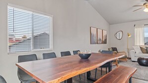Een moderne eetruimte met een houten tafel, zwarte stoelen en een bankje. Drie kunstwerken in lijsten hangen aan de muur, er staat een vloerlamp en er is uitzicht op het buitenlands gebied door grote ramen.