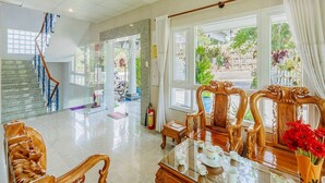A spacious living area with a staircase, a dining table set with cups and a teapot, and a view of the outdoors through large windows. - Capital O 1207 Green Hill Villa Da Lat (Xuan Truong - Da Lat)