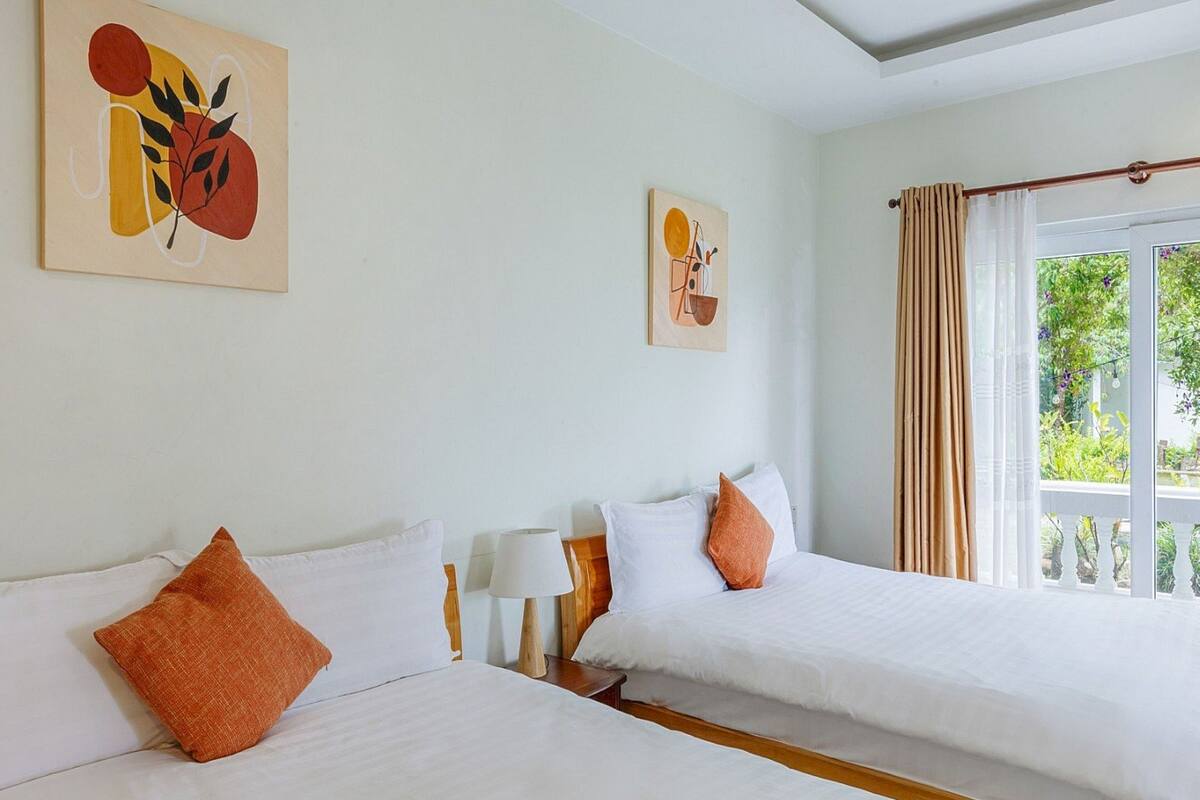 Habitación de hotel con dos camas, ropa de cama blanca, almohadas anaranjadas, cabecera de madera, mesita de noche con lámpara y obras de arte en las paredes.