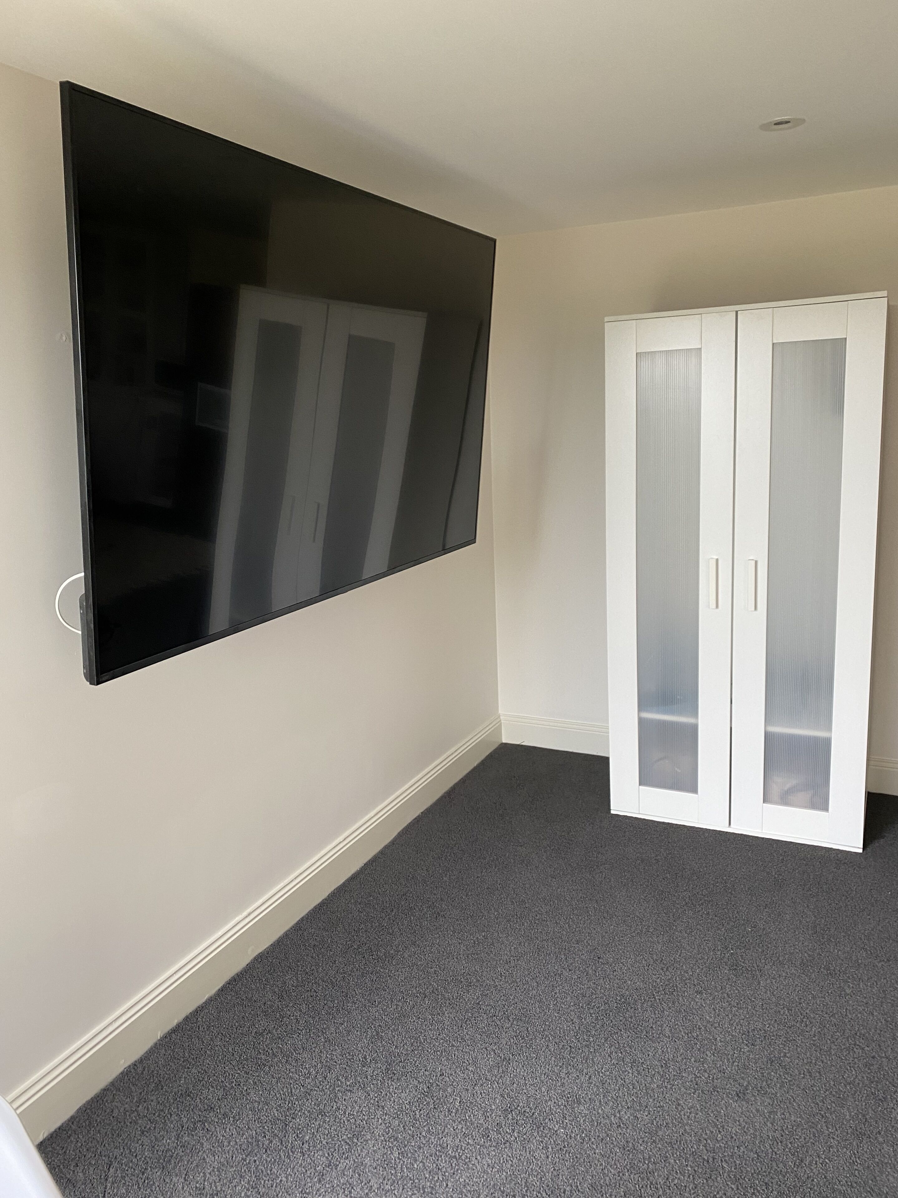A room with a large flat-screen TV mounted on the wall, a white wardrobe with glass doors, and a grey carpeted floor.