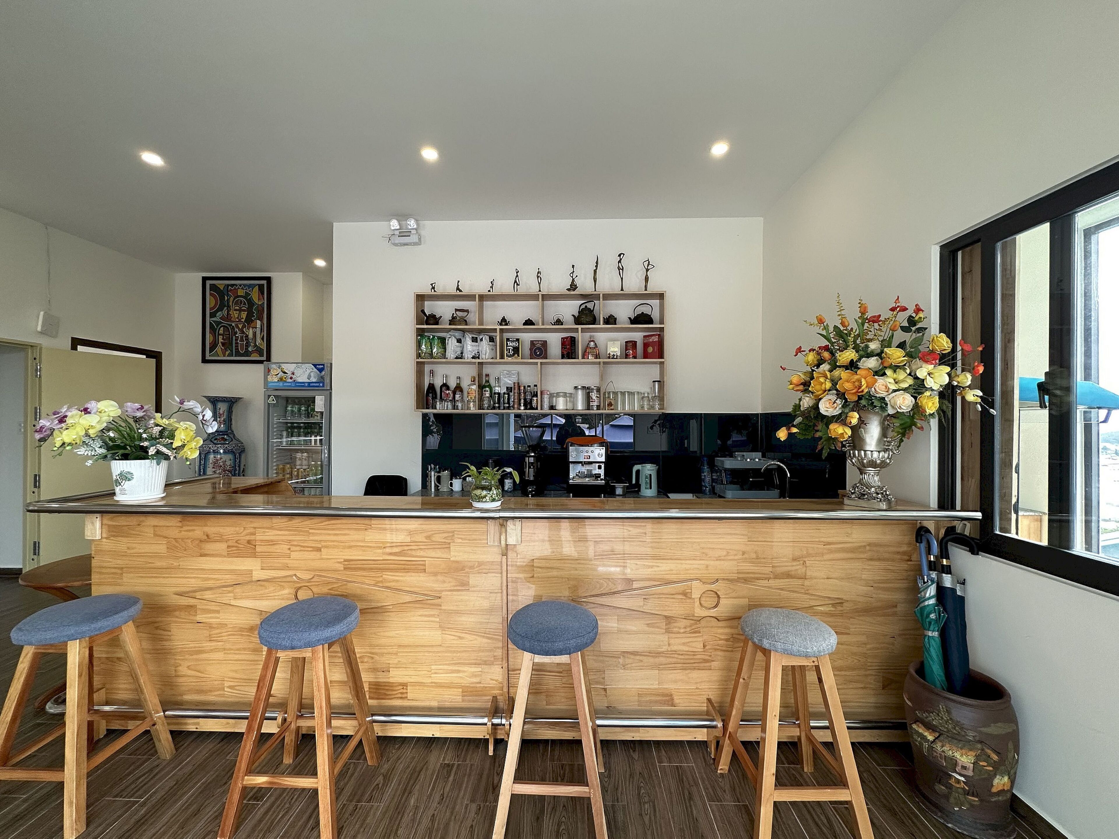 Zona de bar con mostrador de madera, tres taburetes, una estantería con botellas y vasos, y un amplio ventanal.