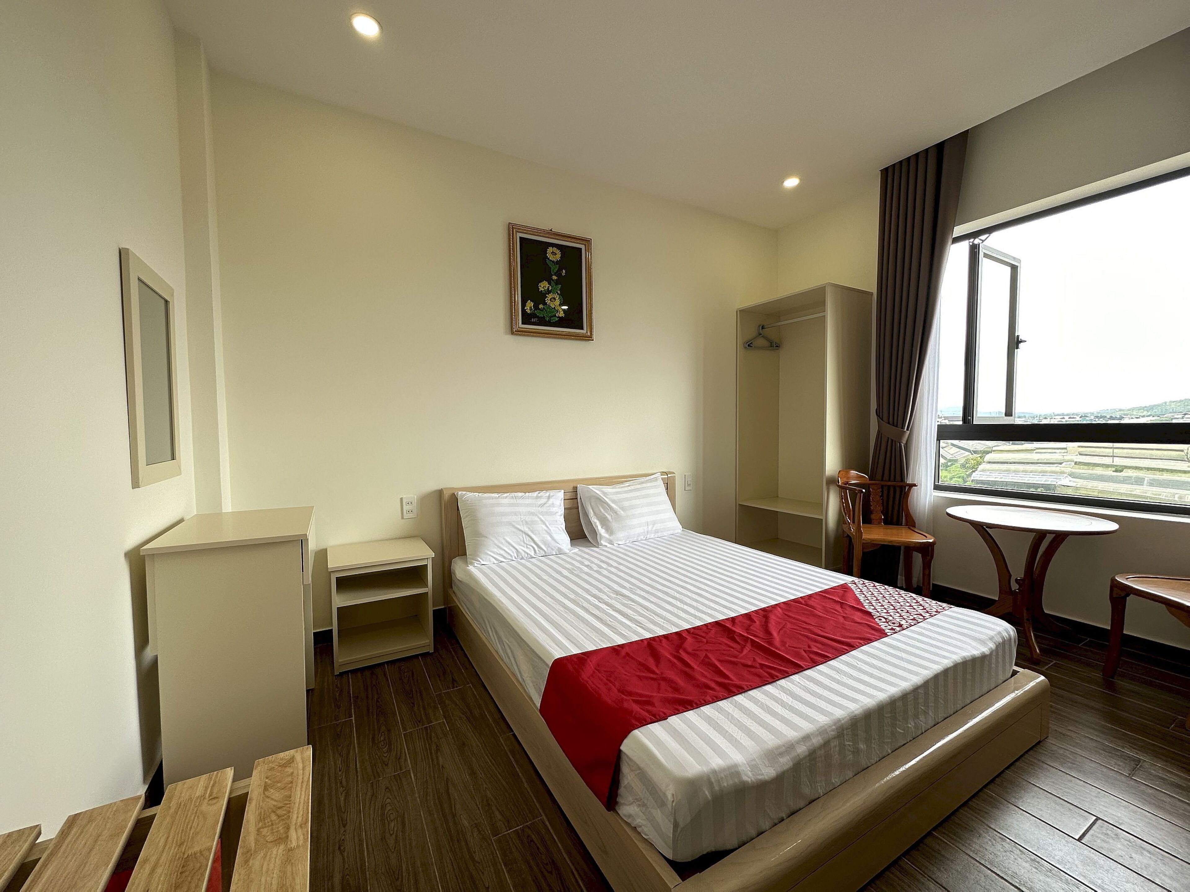A hotel room with a large bed, a bedside table, a small table, a chair, a wardrobe, and a window with a view.