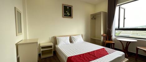 A hotel room with a large bed, a bedside table, a small table, a chair, a wardrobe, and a window with a view.