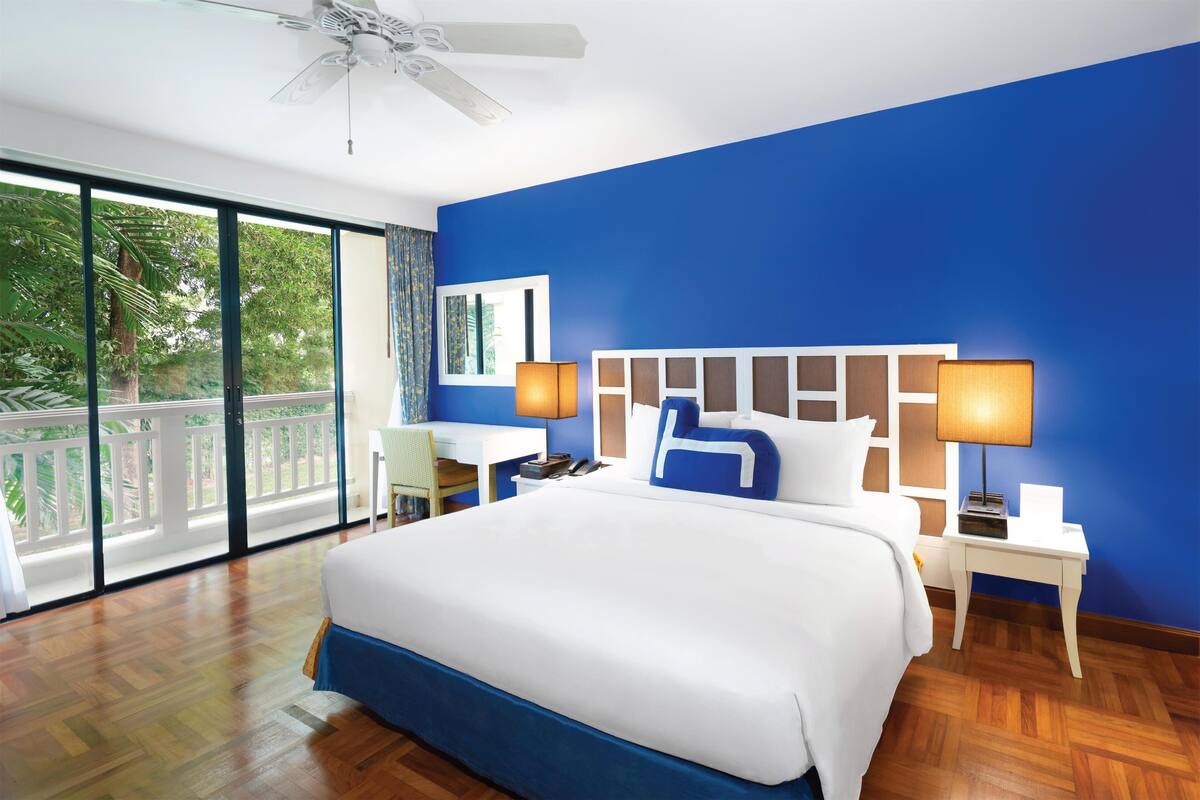 A modern hotel room with a large bed, a desk, and a balcony with a view of greenery.