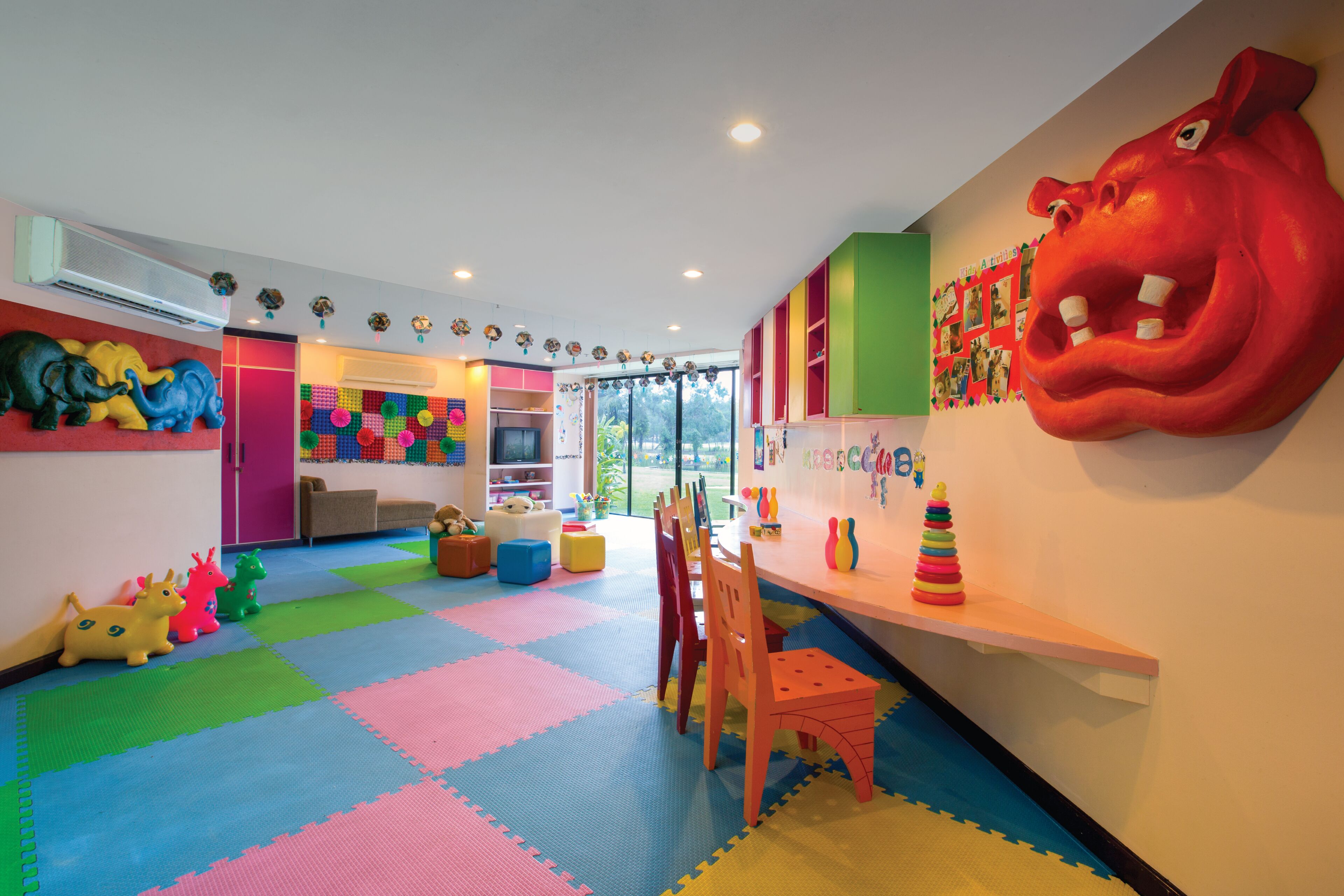 A brightly colored playroom with a large red hippopotamus mural, colorful play mats, and various toys.