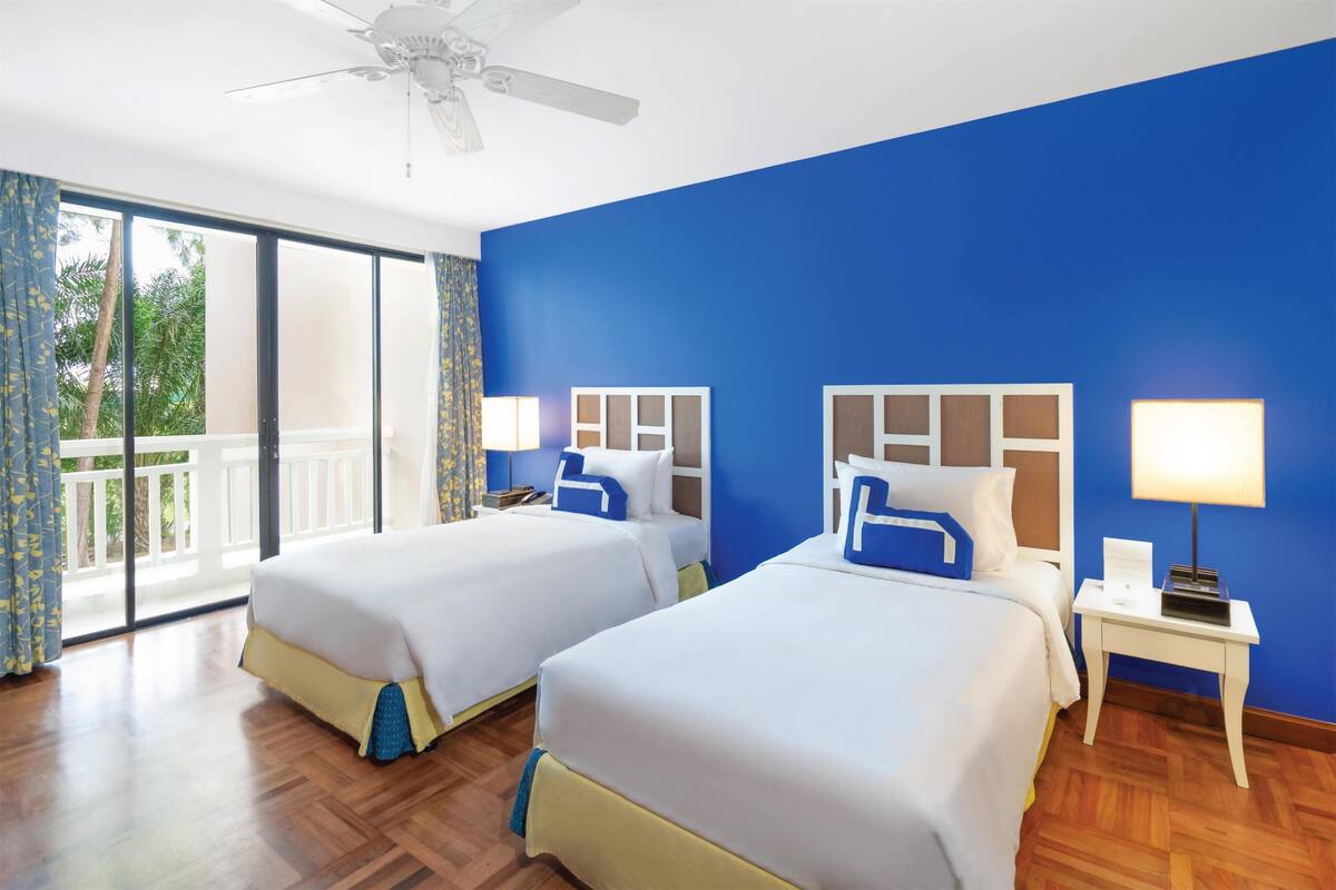 A hotel room with two beds, a ceiling fan, a balcony with curtains, and a nightstand with a lamp.