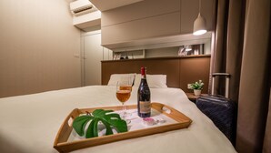 A hotel room with a bed, a bedside table with a bottle of wine, two glasses of wine, a tray with a plant, and a suitcase.