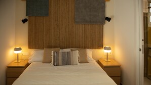 Una cama bien hecha con dos lámparas de noche, un cabecero con textura tejida y una puerta que da a un baño.