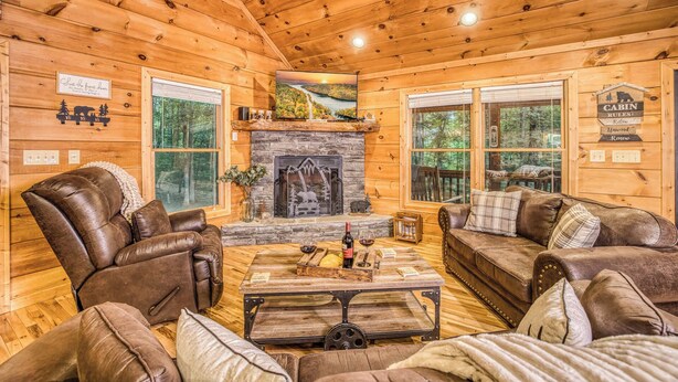 A cabin interior with a stone fireplace, leather sofas, a coffee table, and a television mounted above the fireplace.