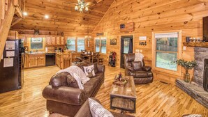 A log cabin interior with a kitchen, dining area, and living room furnished with leather sofas and a fireplace.