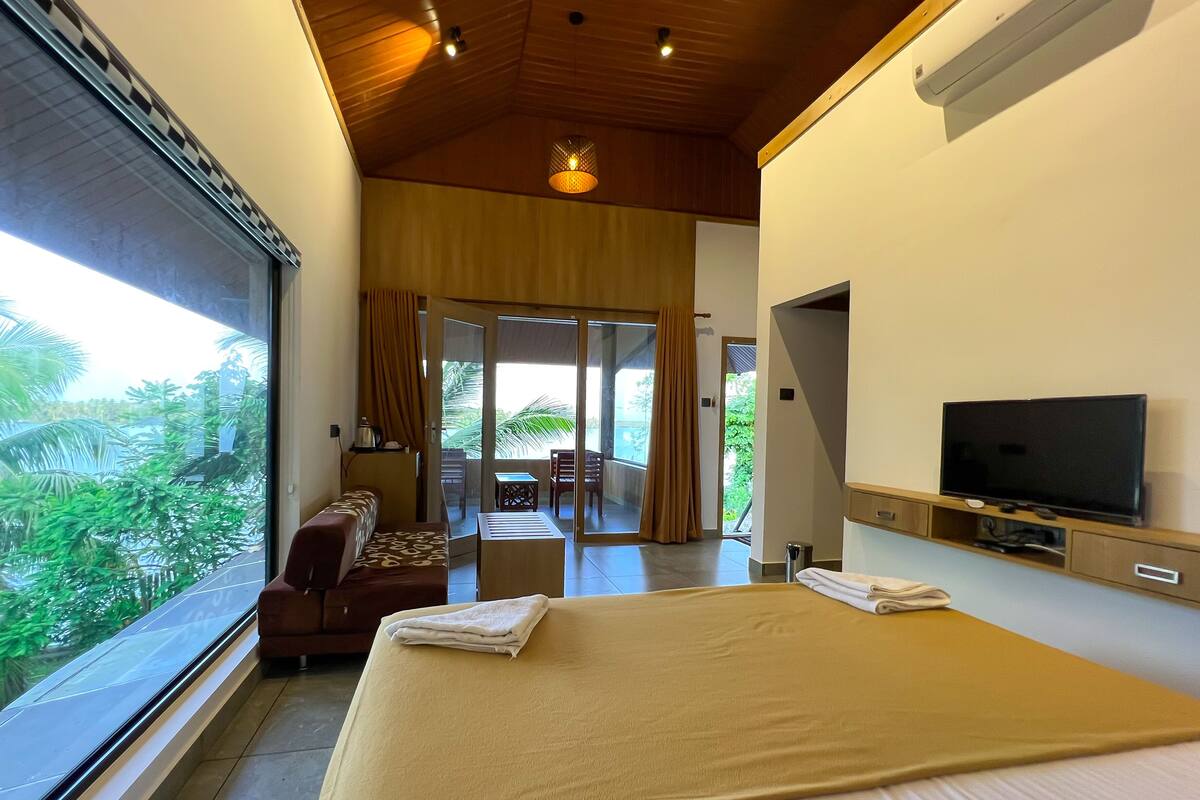 A hotel room with a large bed, a TV, a sofa, and a balcony with a view of greenery.
