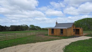 A wooden cabin with a gravel driveway leading to it, surrounded by a fenced pasture and trees. - Renagour Cottage - Rural charm with outdoor hot tub! (Mangamahu)