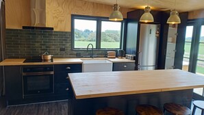 A modern kitchen with a large island, bar stools, a built-in oven, and a view of the outdoors through a window. - Renagour Cottage - Rural charm with outdoor hot tub! (Mangamahu)