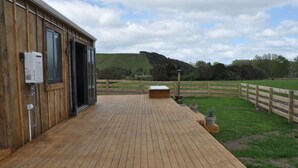 A wooden deck leading to a building with a large window and an open door. - Renagour Cottage - Rural charm with outdoor hot tub! (Mangamahu)