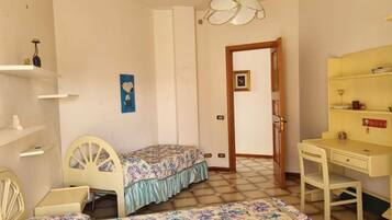 A bedroom with a bed, a desk, shelves, a chair, and a door leading to another room.