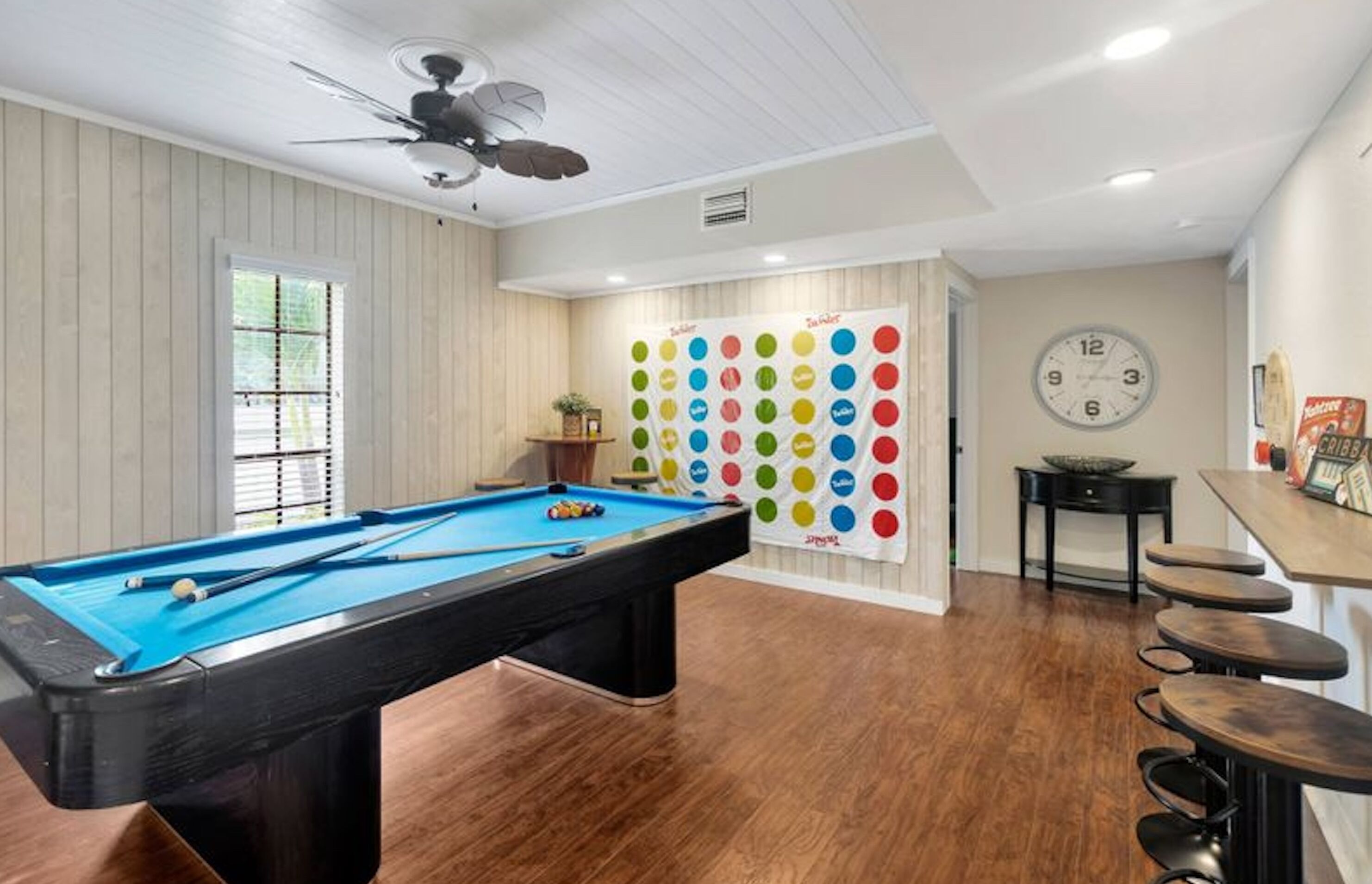 A pool table in a room with wooden flooring, a ceiling fan, and a wall clock.