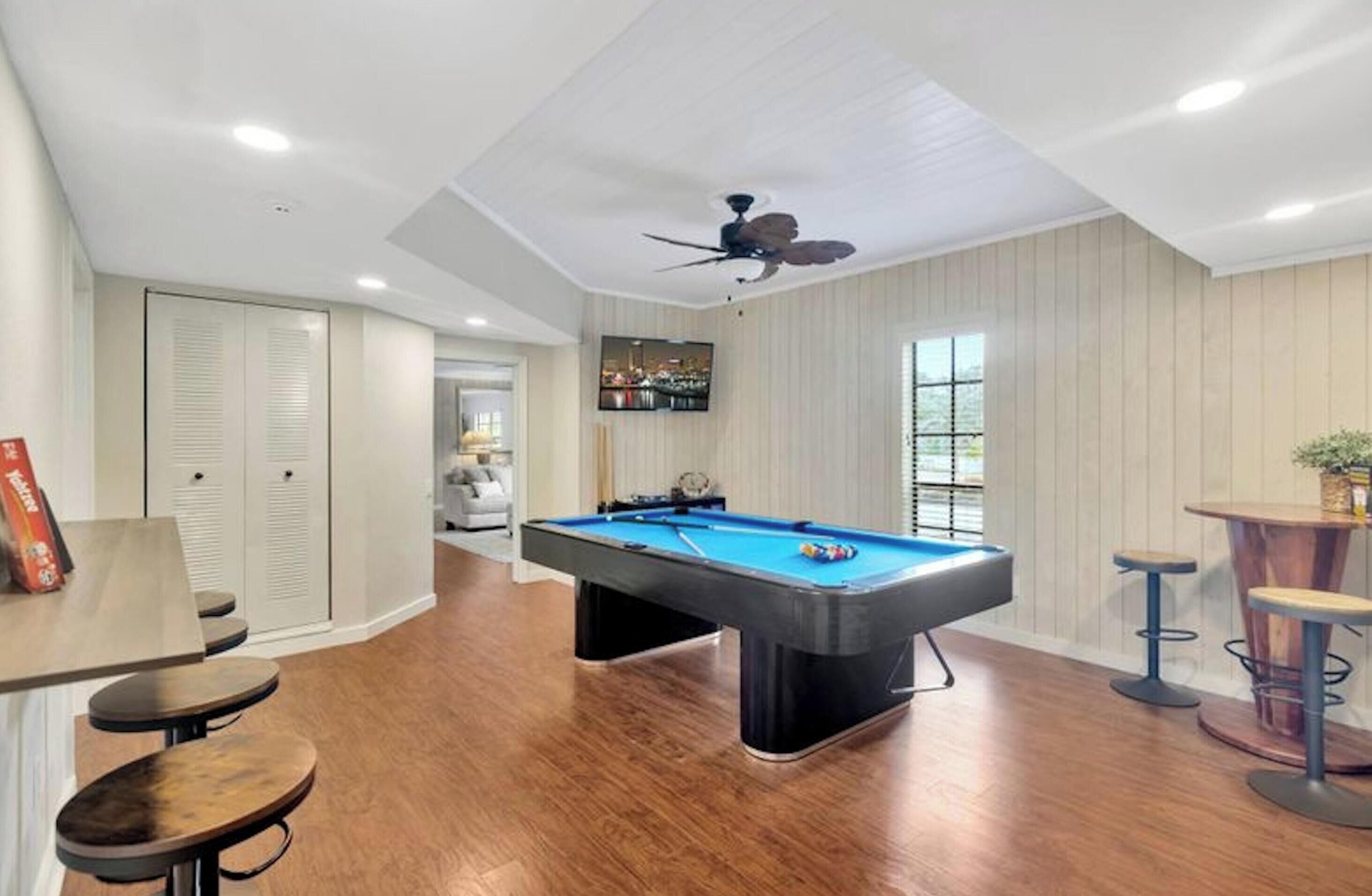 Une table de billard dans une pièce au plancher en bois, avec un ventilateur de plafond et une fenêtre équipée de stores.