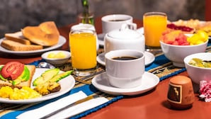 Una mesa de desayuno con tostadas, huevos, aguacate y fruta. Hay tazas de café y jugo de naranja.
