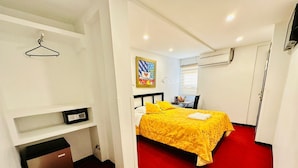 Una habitación de hotel con una cama, un escritorio con una computadora, un perchero y un cuadro en la pared, además de un equipo de aire acondicionado.
