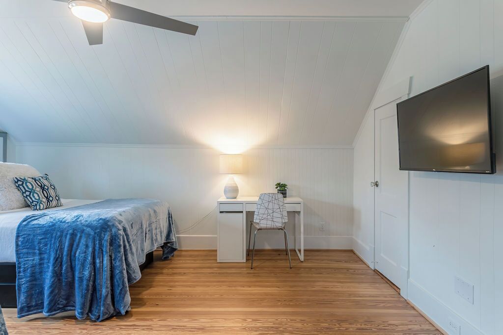 Ein Schlafzimmer mit einem Bett, einem Schreibtisch mit Stuhl, einem an der Wand befestigten Fernseher und einem Deckenventilator.