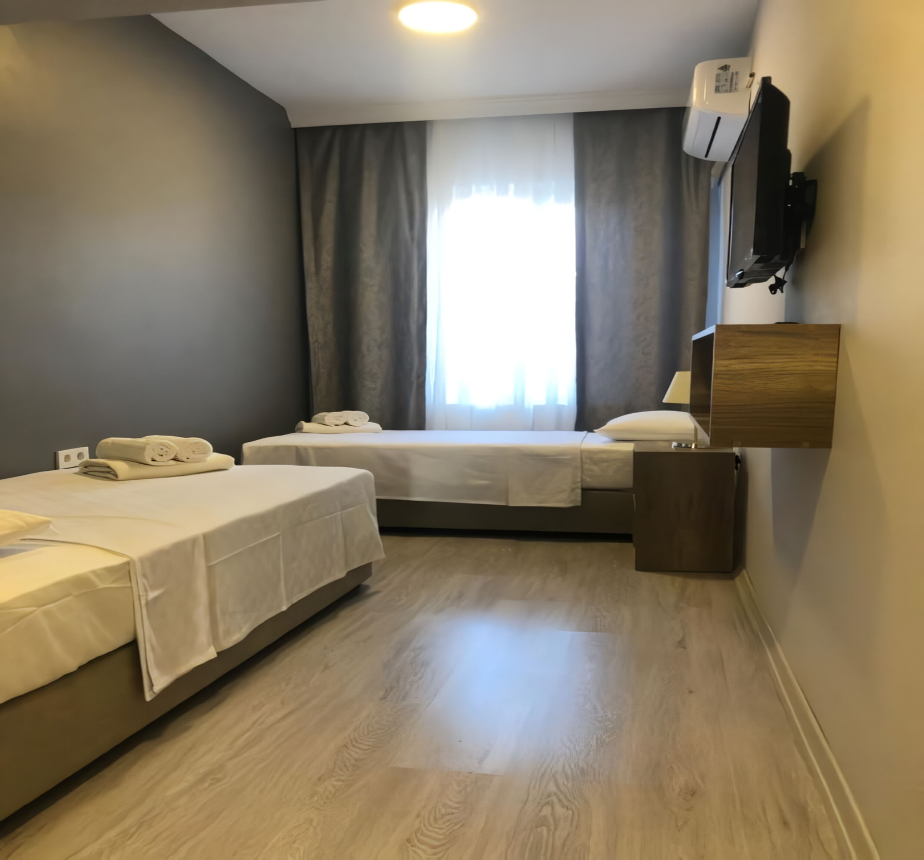 A small, modern hotel room with two beds, a wooden floor, a window with curtains, a mounted TV, and a wall-mounted air conditioner.