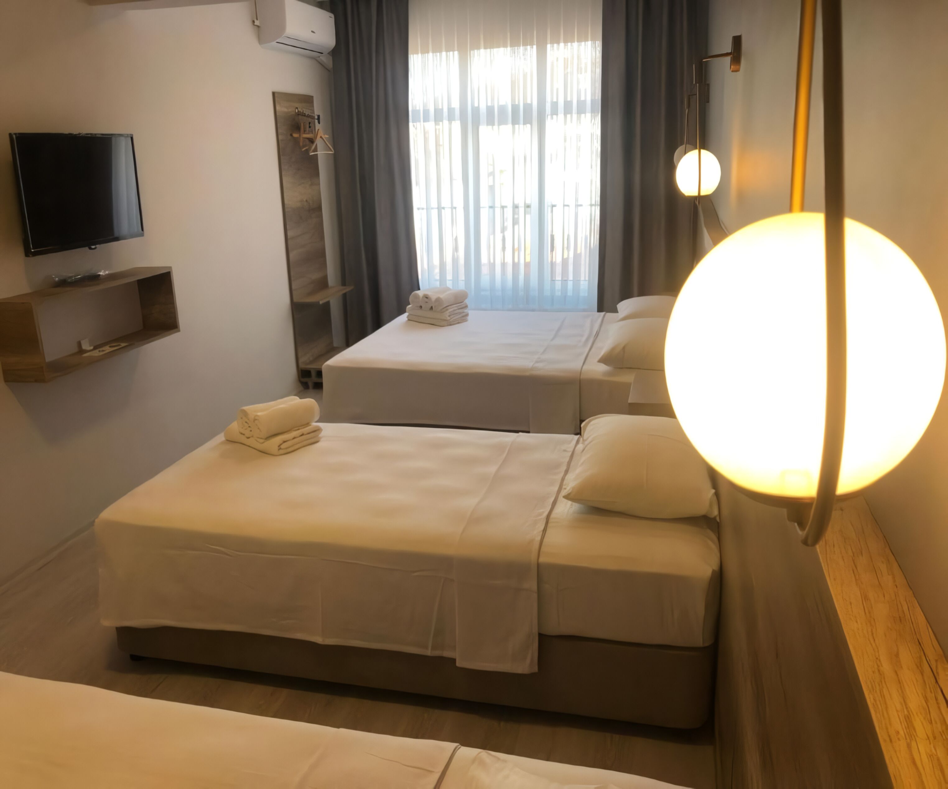 Habitación de hotel con dos camas, televisión, repisa con toallas y una lámpara redonda grande.