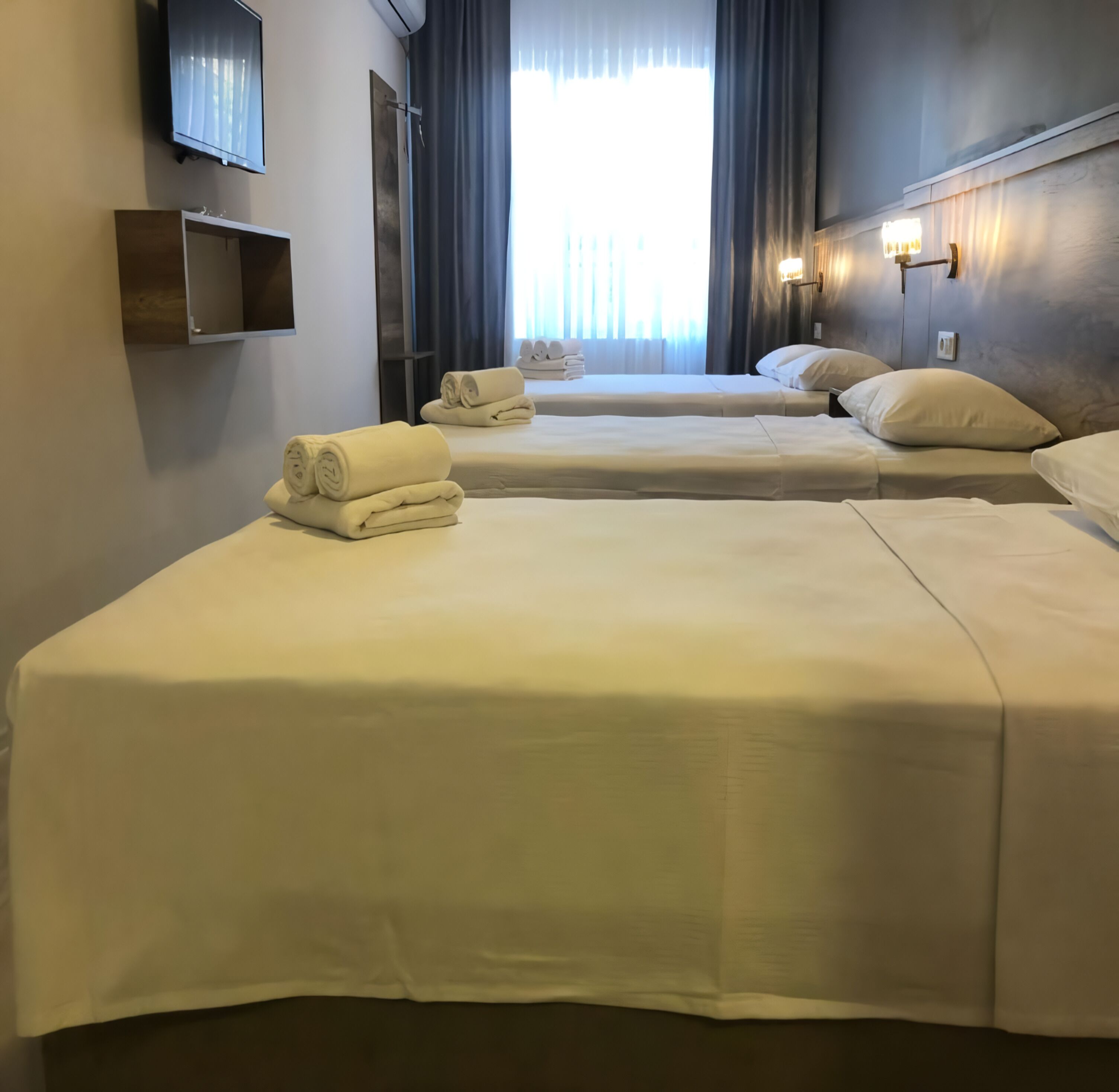 A hotel room with two beds, each with neatly folded towels, a flat-screen TV mounted on the wall, and a bedside lamp.