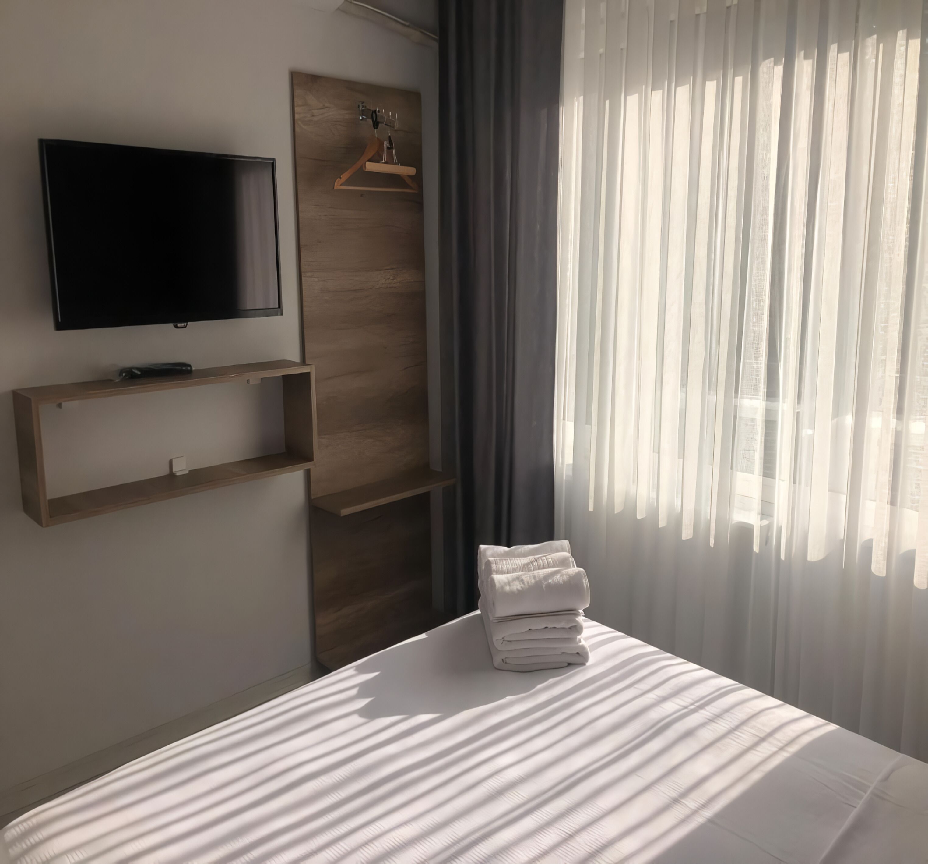 Une chambre d’hôtel avec un lit, un téléviseur fixé au mur, un panneau en bois, un porte-vêtements et des rideaux légers.