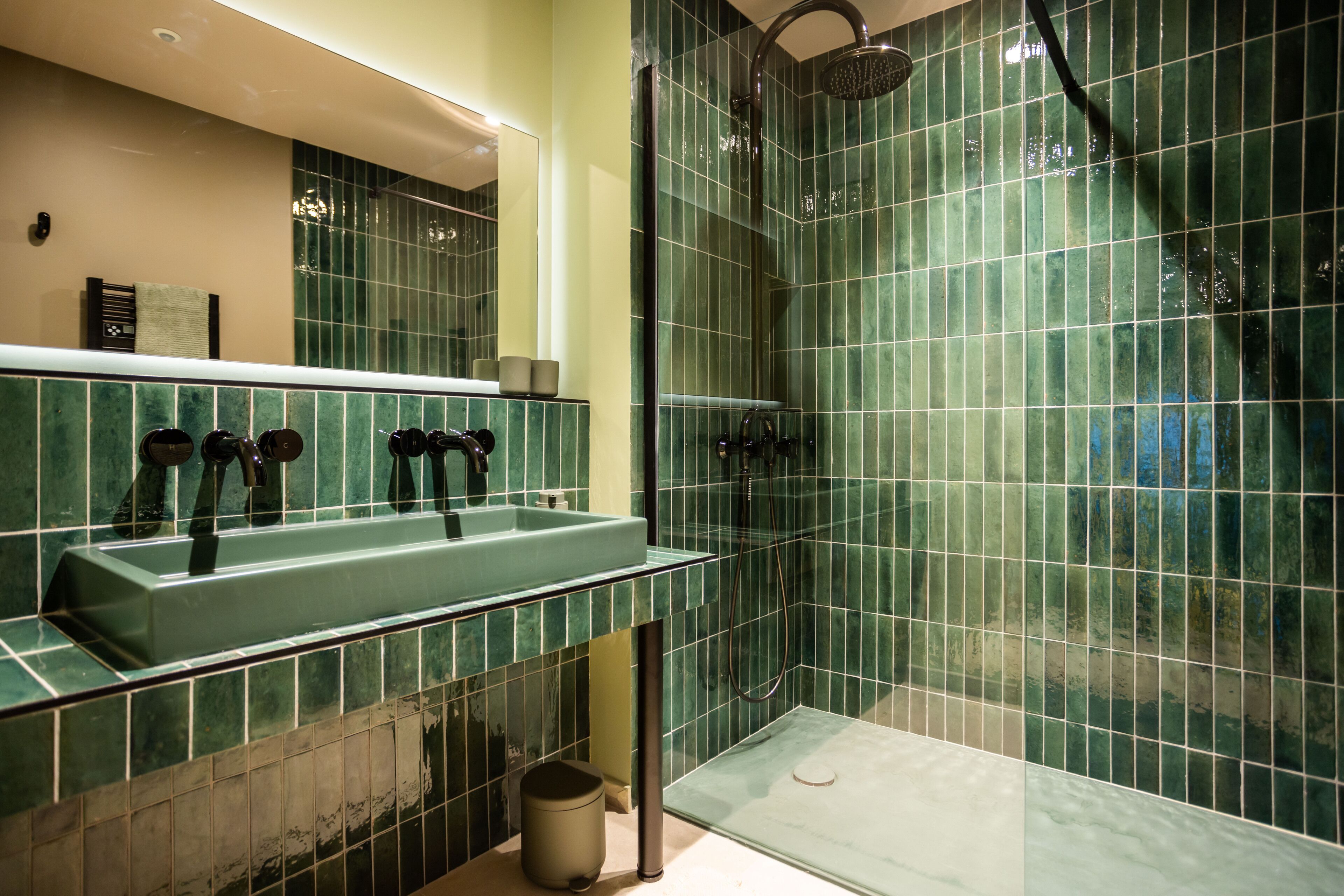 Une salle de bain avec une douche carrelée de carreaux verts, un lavabo avec un miroir au-dessus et une poubelle.