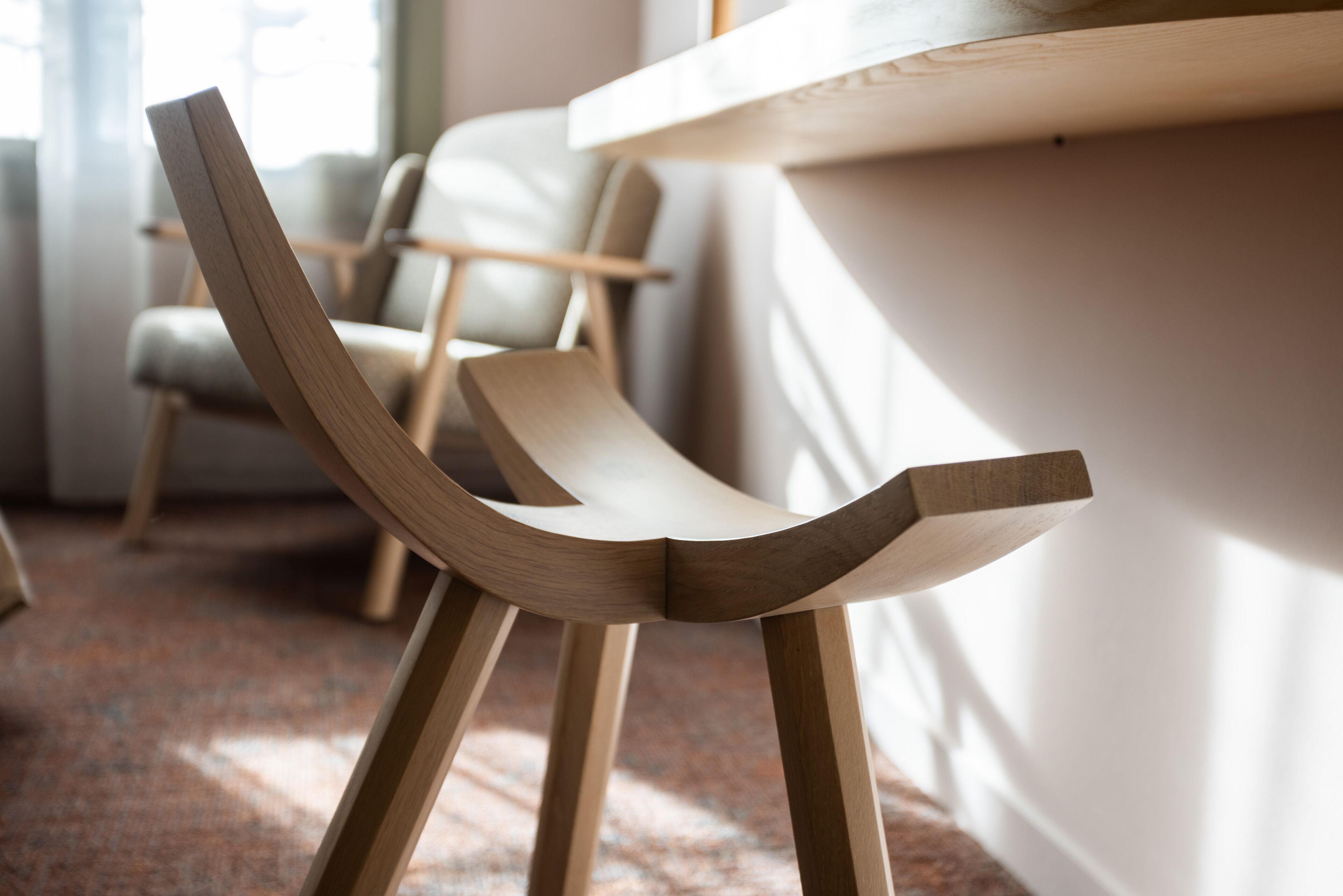 A modern wooden chair with a unique curved backrest design, positioned in a well-lit room with a carpeted floor and a white wall in the background.