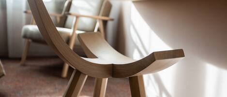 A modern wooden chair with a unique curved backrest design, positioned in a well-lit room with a carpeted floor and a white wall in the background.