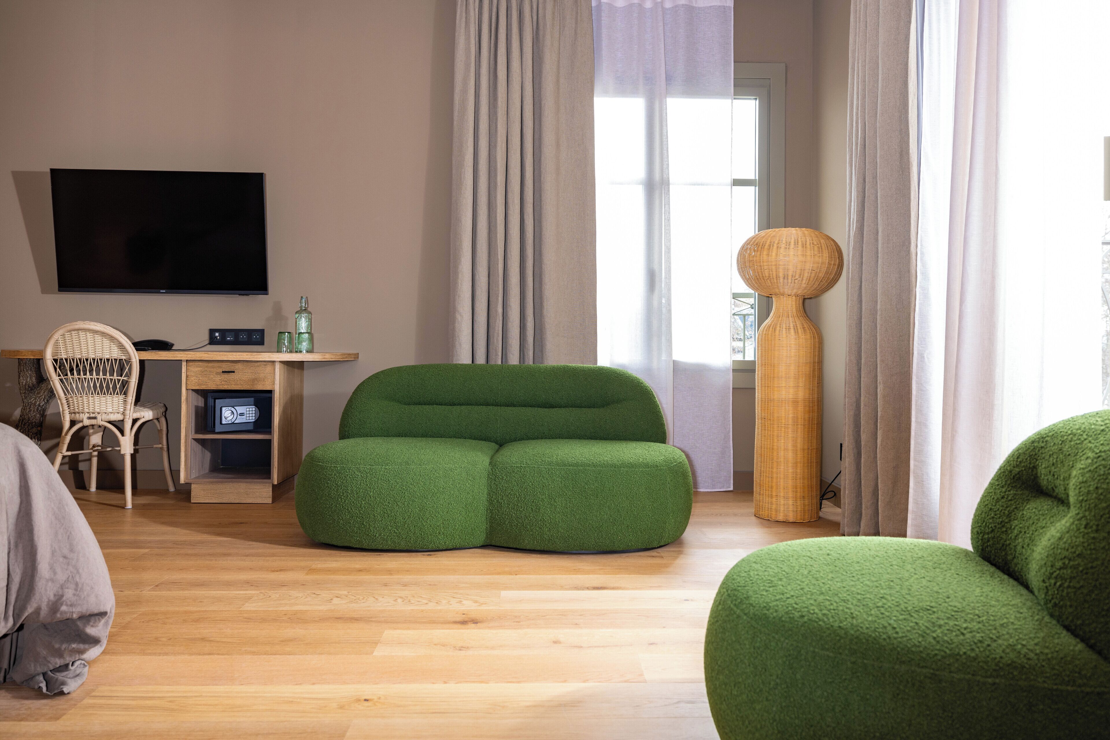 A modern living room with a green sofa, a wooden desk with a flat-screen TV, a wicker chair, and a tall woven lamp.