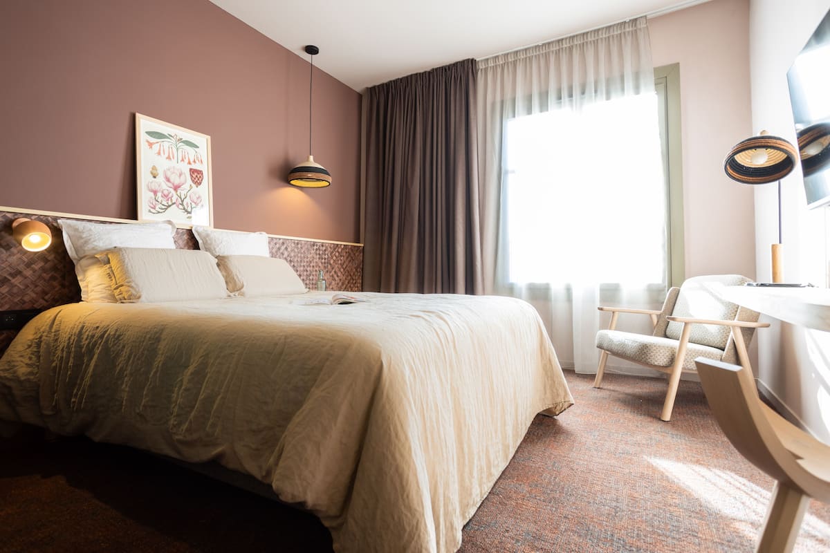 Un lit bien fait, avec une couette et des oreillers de couleur beige, un bureau en bois avec une chaise, une grande fenêtre avec des rideaux et un luminaire fixé au mur.