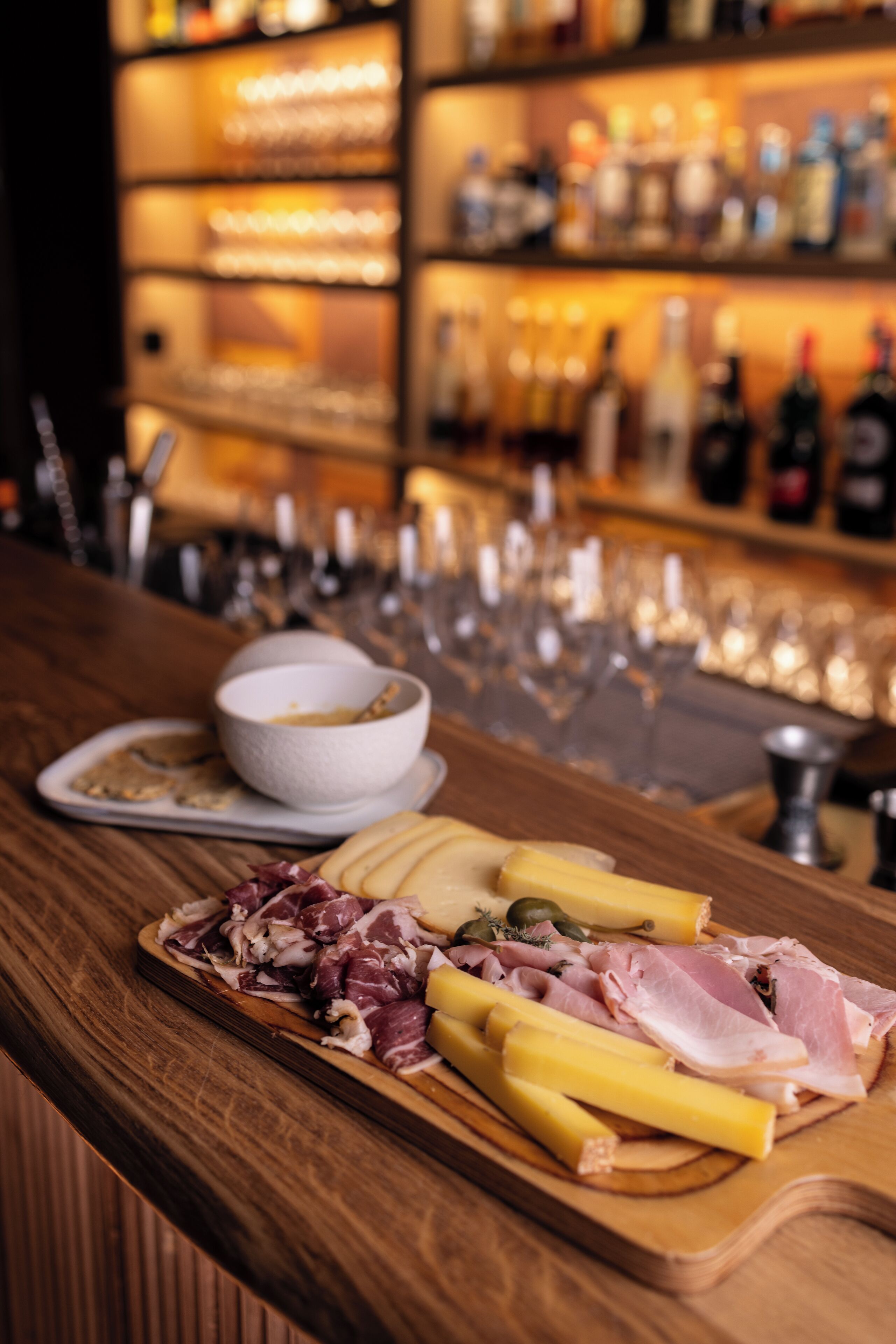 Una zona de bar con mostrador de madera mostrando una variedad de quesos, embutidos y aceitunas.