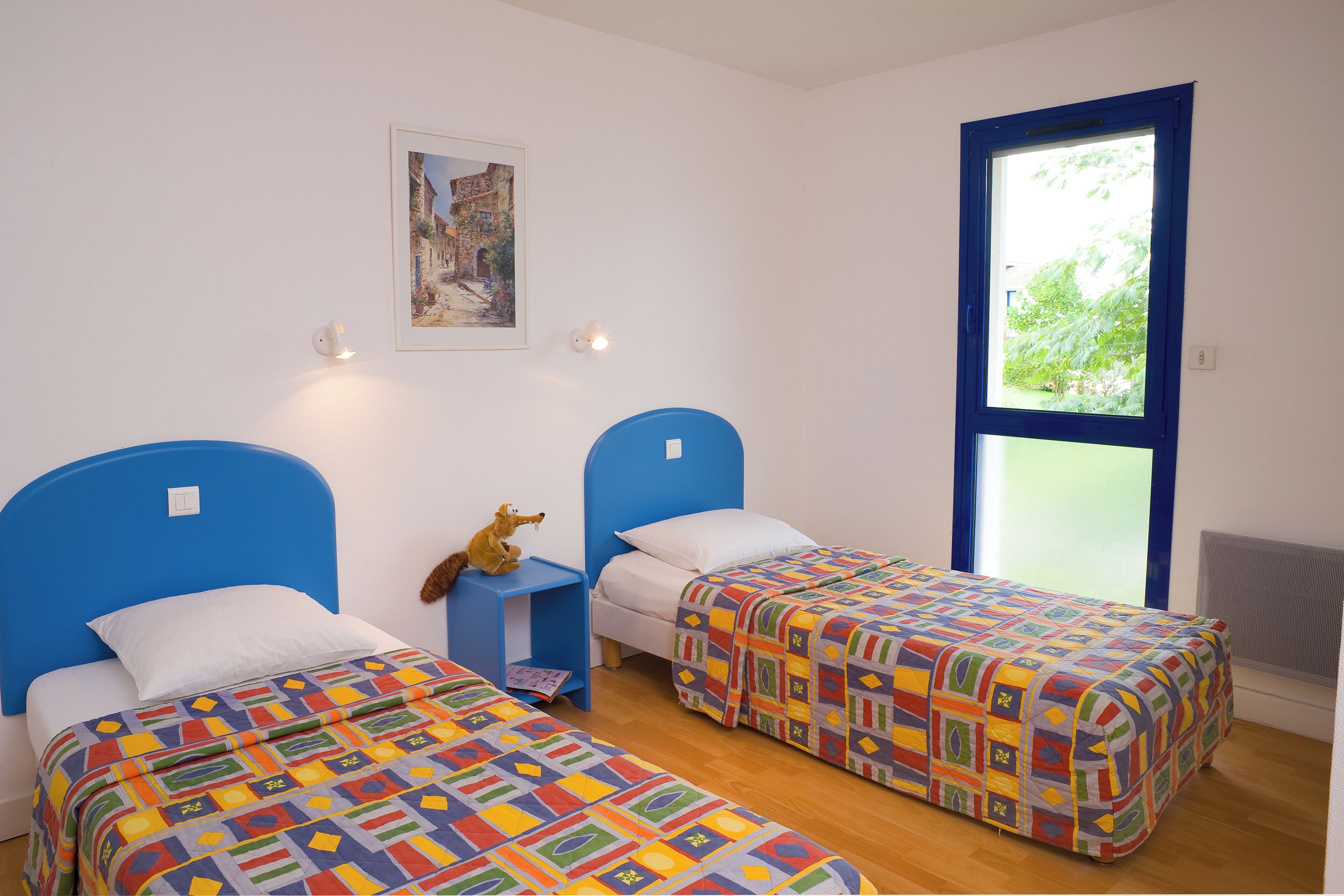 A room with two beds, a small table, a wall-mounted light, and a window with a view of greenery.
