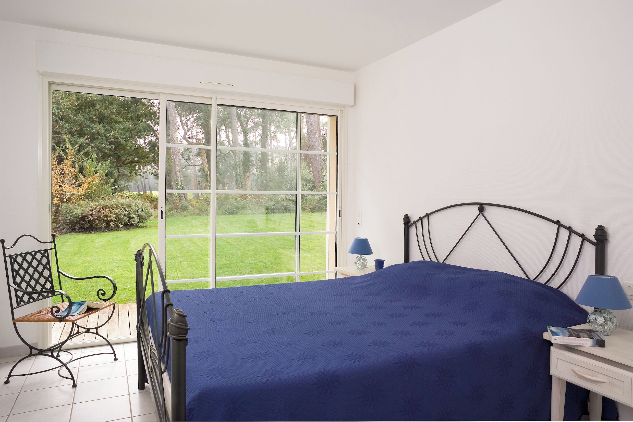 A bedroom with a large window, a bed, a chair, a nightstand, and a lamp.