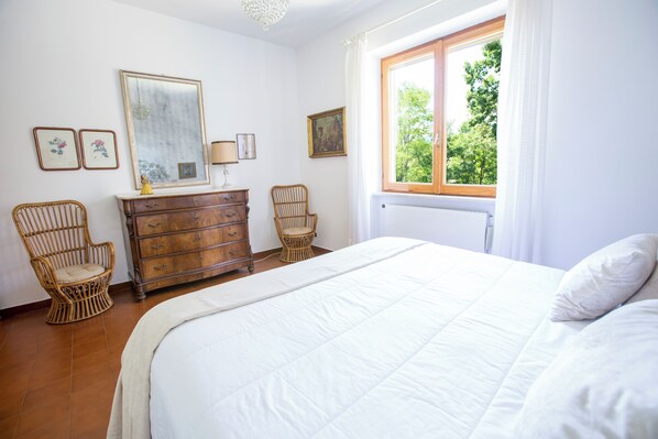 A bedroom with a large bed, two wicker chairs, a wooden dresser, and a window with a view of trees.
