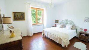 Une chambre à coucher avec un lit, une table de chevet, une commode, une fenêtre avec des rideaux, un lustre et un tableau au mur.