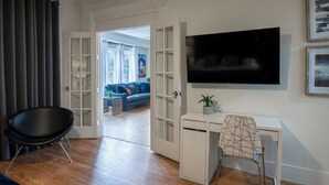 Een moderne woonkamer met een flatscreen televisie aan de muur, een wit bureau met een stoel, een zwarte stoel en een deur naar een andere kamer.